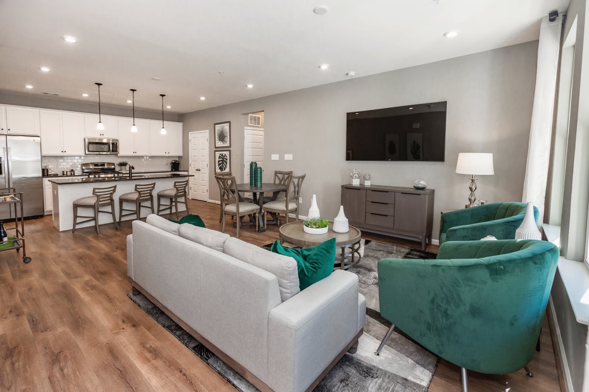Open floor plan in the Lincoln by HHHunt Homes at The Pointe at Twin Hickory, featuring a modern kitchen with white cabinetry and stainless steel appliances, a central island with bar seating, and a cozy living area with green accent chairs and a wall-mou