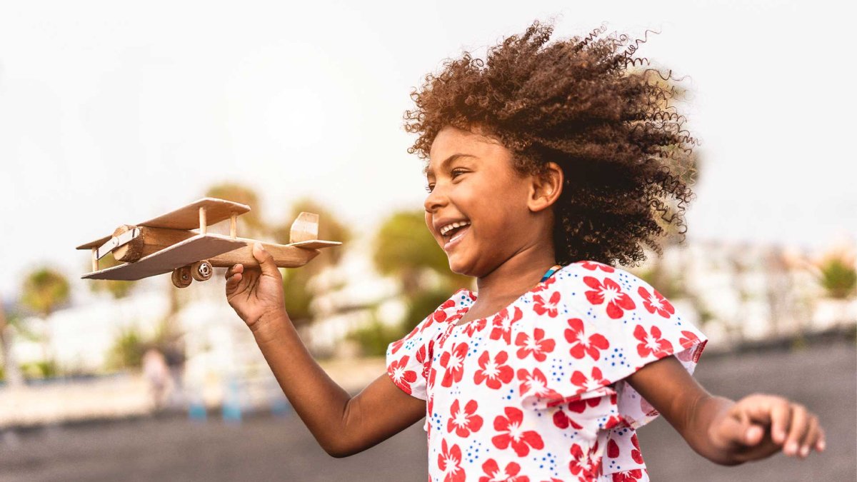 A young running outside and smiling. She holds up a wooden airplane in the air in front of her.