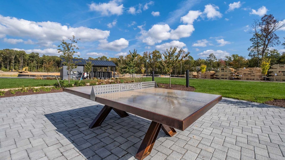 Outdoor ping pong table at Wescott by HHHunt, set on a paved patio with landscaped green space and community features in the background under a blue sky.