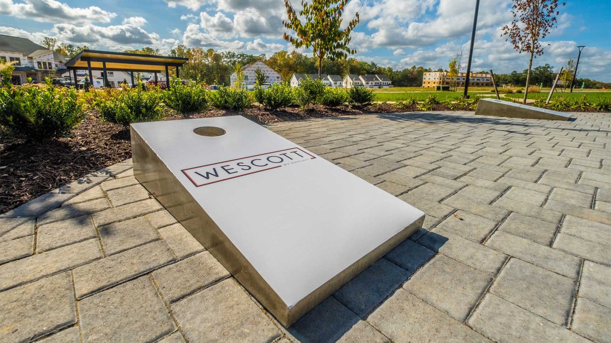 Outdoor cornhole boards at Wescott by HHHunt, set on a paved patio with landscaped green space and community features in the background under a blue sky.