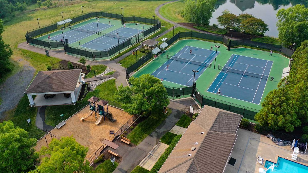 Amenity area at The Pointe at Twin Hickory by HHHunt Homes, featuring multiple lighted tennis courts, a fenced playground with slides, a picnic pavilion, and nearby pool.