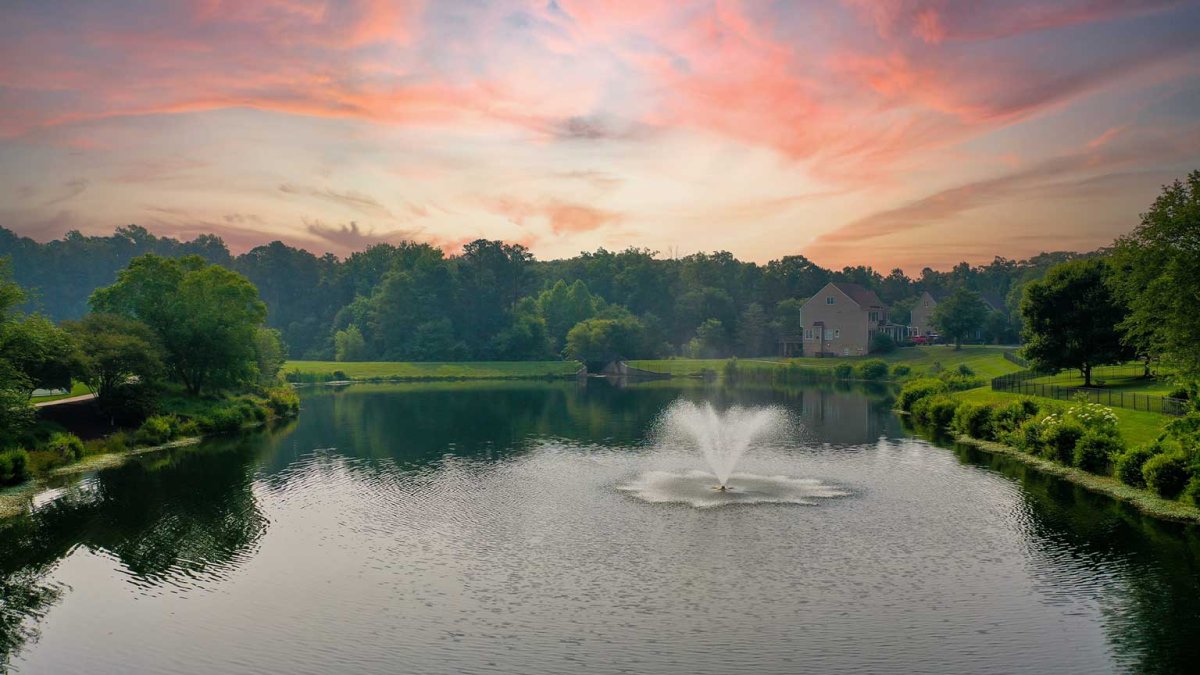Sunset over the lake with fountain at The Pointe at Twin Hickory by HHHunt, surrounded by trees and nearby homes, highlighting tranquil community views.