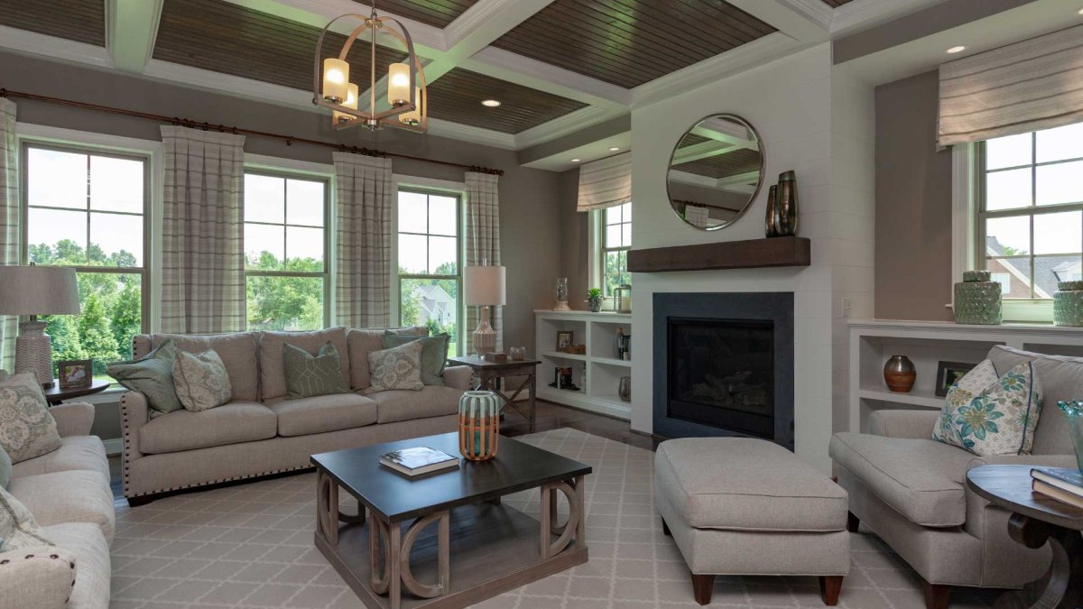 Living room in the Crawford floor plan by HHHunt Homes at The Summit At Morgan Ridge, featuring a coffered ceiling with stained wood insets, a white shiplap fireplace with dark wood mantel, built-in shelving, multiple large windows with plaid curtains, an