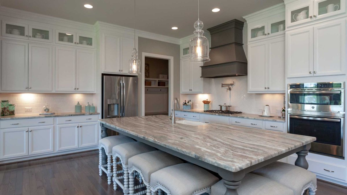 Kitchen in the Crawford floor plan by HHHunt Homes at The Summit At Morgan Ridge featuring a large marble-look island with spindle-leg stools, white cabinetry with glass accents, stainless steel appliances, a custom range hood, pendant lighting, and a pot