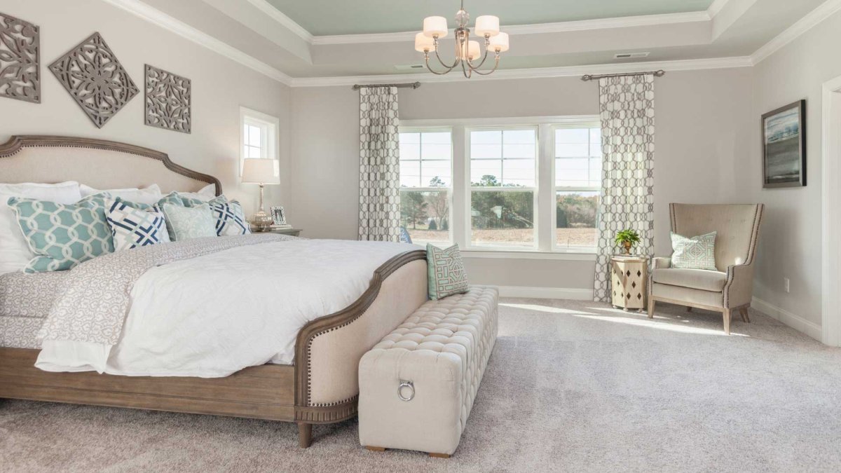 Bedroom in an HHHunt Home at The Summit At Morgan Ridge featuring a tray ceiling with crown molding, large windows with patterned drapes, a tufted bench at the foot of a wood-framed bed, coordinating accent pillows, neutral carpet, and a cozy reading corn