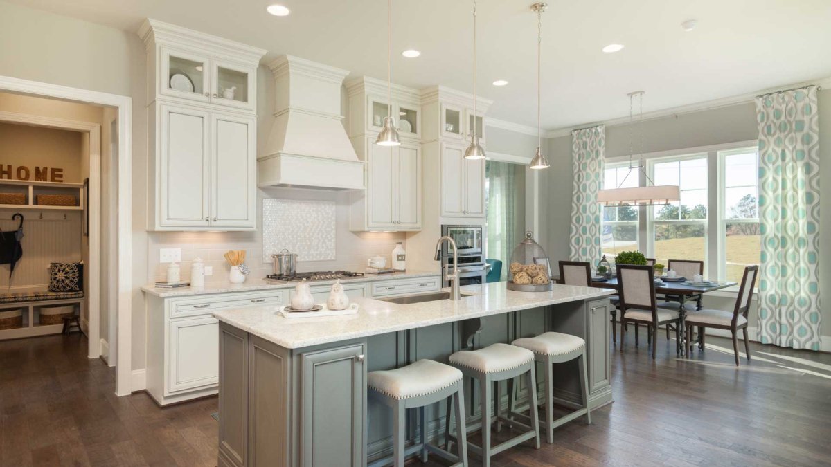 Kitchen by HHHunt Homes at The Summit At Morgan Ridge, featuring a large central island with seating, stainless steel appliances, gas cooktop, white cabinetry with glass inserts, and an adjacent dining area with natural light from tall windows and pattern