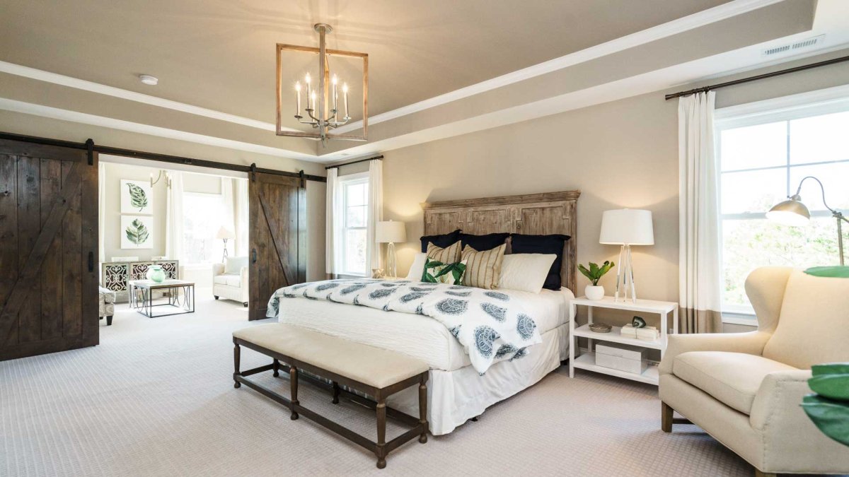 Primary bedroom in an HHHunt Home at The Summit At Morgan Ridge featuring a tray ceiling with crown molding, a rustic wood headboard, neutral-toned decor, and sliding barn doors leading to a sitting area with natural light.
