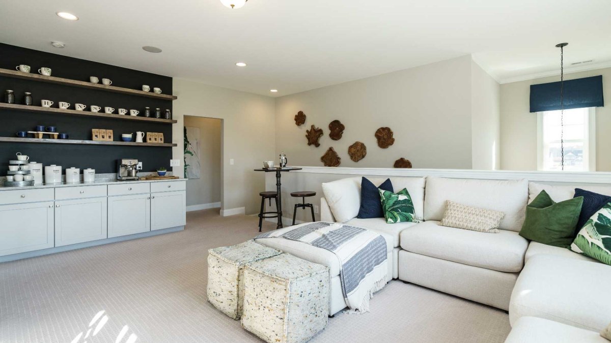 Loft in an HHHunt Home at The Summit At Morgan Ridge, featuring a built-in coffee bar with open shelving, modern light gray cabinetry, a cozy sectional sofa with green and navy accent pillows, soft carpet flooring, and a bistro-style table for casual seat