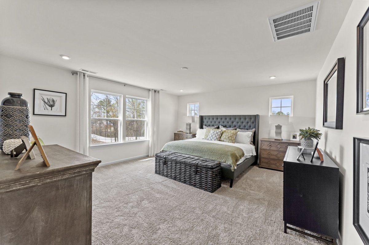 Primary bedroom in the Curie floor plan at Qualla Trace by HHHunt Homes, showcasing a tufted headboard, large windows, soft carpet flooring, and natural light.