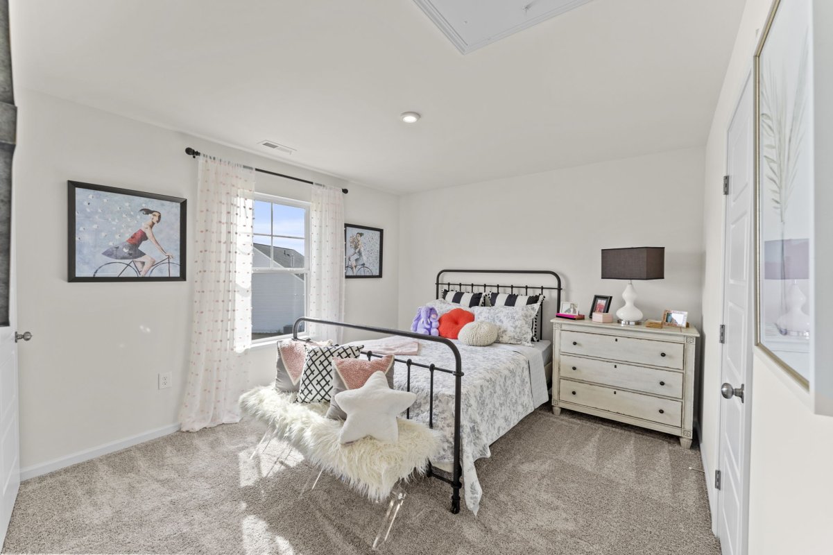 Secondary bedroom in the Qualla Trace community by HHHunt Homes, featuring a black metal bed frame, soft carpet flooring, large window with sheer curtains, and neutral décor with playful accents.