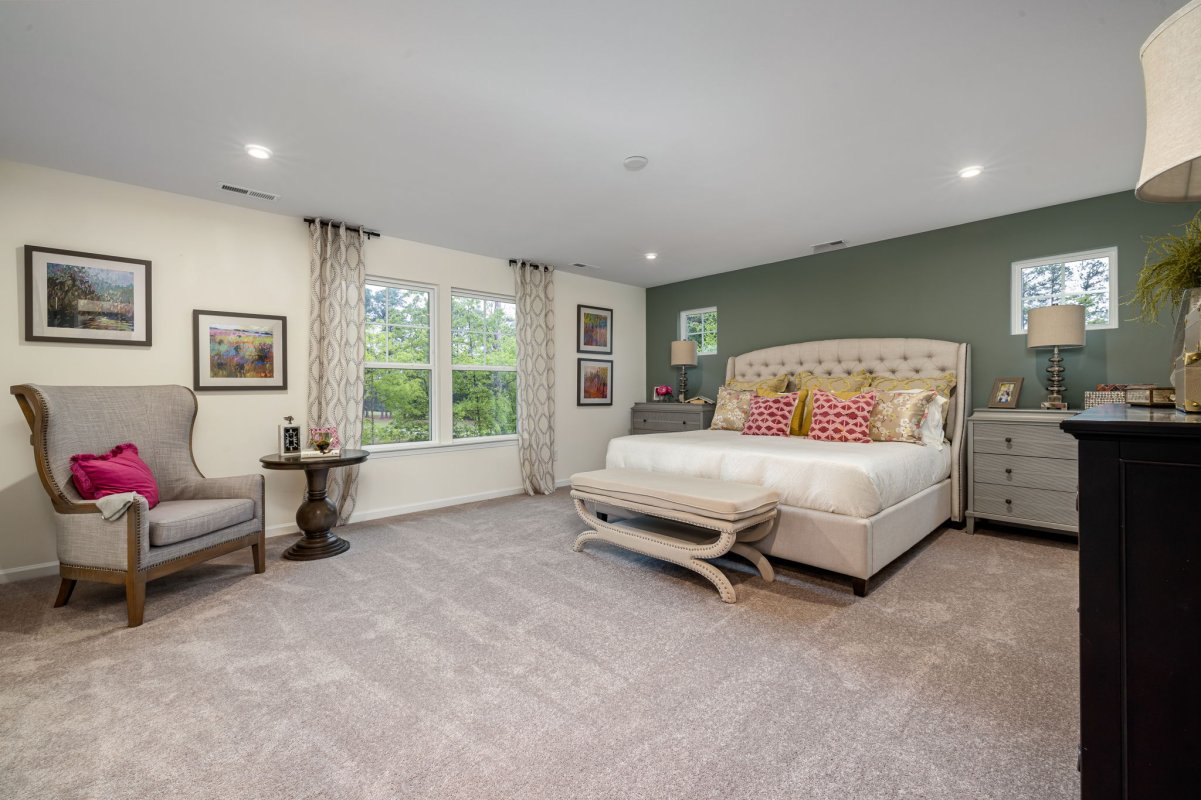 Primary suite in the Edison floor plan by HHHunt Homes at Qualla Trace, featuring a tufted upholstered bed, green accent wall, neutral carpeting, and large windows with patterned curtains and natural light.