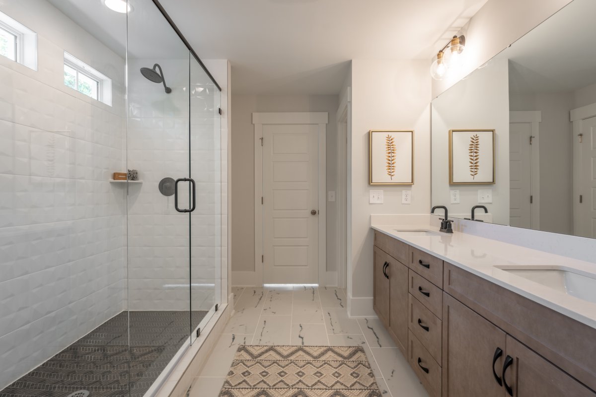 Primary bathroom in the Grayson floor plan at Tyson’s Trail Estates by HHHunt Homes, featuring a frameless glass walk-in shower with textured white tile, double vanity with light wood cabinetry, quartz countertops, and modern black fixtures.