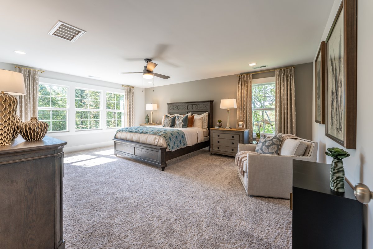 Spacious primary bedroom in the Grayson floor plan at Tyson’s Trail Estates by HHHunt Homes, featuring large windows with abundant natural light, neutral-toned carpet, a ceiling fan, and a modern gray wood bed frame with matching nightstands and accent de