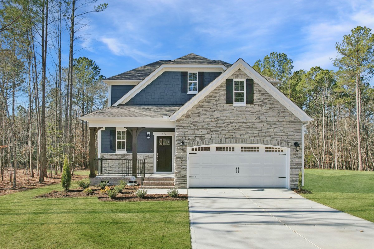 Front exterior view of the Taylor floor plan (European A elevation) by HHHunt Homes at Tyson's Trail Estates, featuring stone siding, a covered porch with dark trim, and a two-car garage surrounded by wooded landscape.