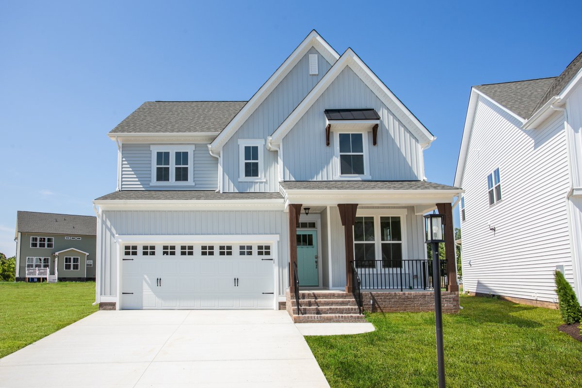 Exterior view of the Morgan floor plan with Farmhouse A elevation at Tyson’s Trail Estates by HHHunt Homes, showcasing a light gray board-and-batten facade, two-car garage, gabled rooflines, and covered front porch with dark wood columns and brick steps.