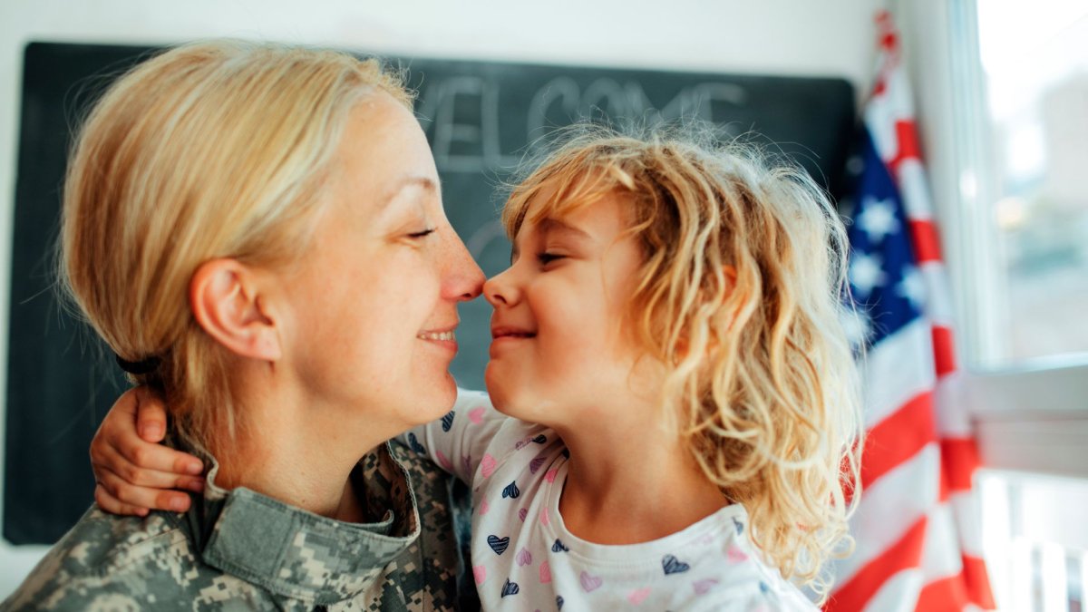 Smiling military mom in uniform embracing her young daughter in front of a welcome home sign and American flag.