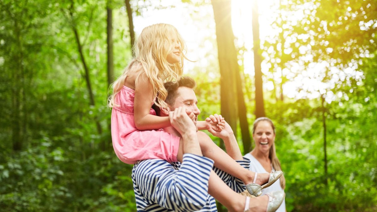 A family of three on a hike through the woods. Dad carries their daughter on his shoulders. They all smile and laugh.