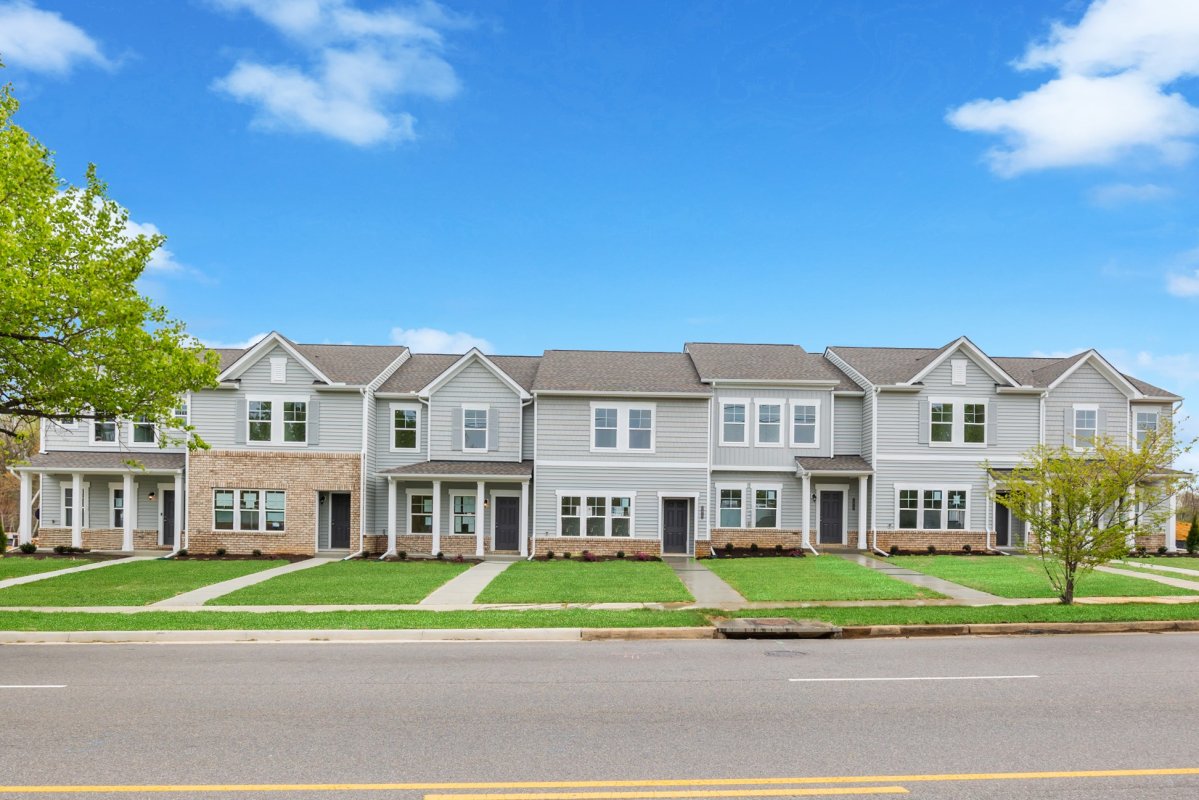 Streetscape view of Kinwick Townhomes by HHHunt Homes, featuring a row of two-story townhomes with gray siding, brick accents, covered entryways, and well-manicured front lawns along a tree-lined street.