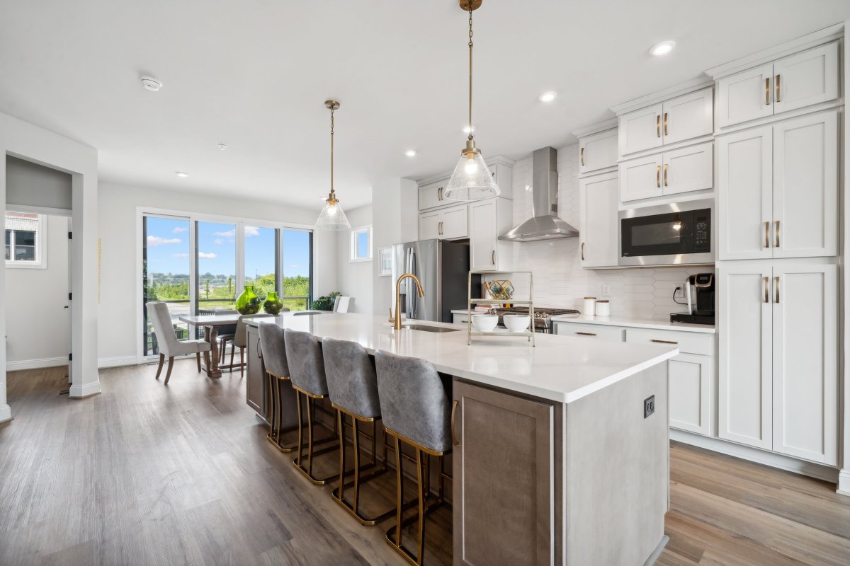 Bright, modern kitchen in the Cameron floor plan at The Aire At Westchester by HHHunt Homes, featuring a large island with quartz countertops, pendant lighting, stainless steel appliances, white shaker cabinets, and wood-style flooring with views of the o
