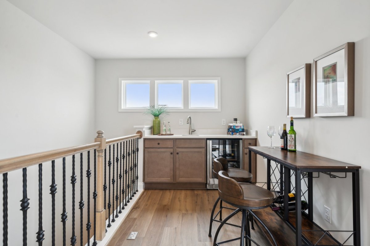 Stylish fourth-floor wet bar in The Aire At Westchester by HHHunt Homes, featuring wood cabinetry, a built-in beverage fridge, stainless steel sink, and a bar-height table with modern stools, set against light walls and large windows with natural light.