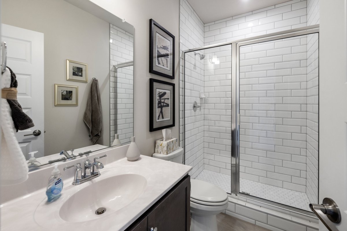 Optional full bathroom in the Drake floor plan at The Aire At Westchester by HHHunt Homes, featuring a white vanity with chrome fixtures, a toilet, and a walk-in shower with white subway tile walls, glass doors, and hexagon tile flooring.