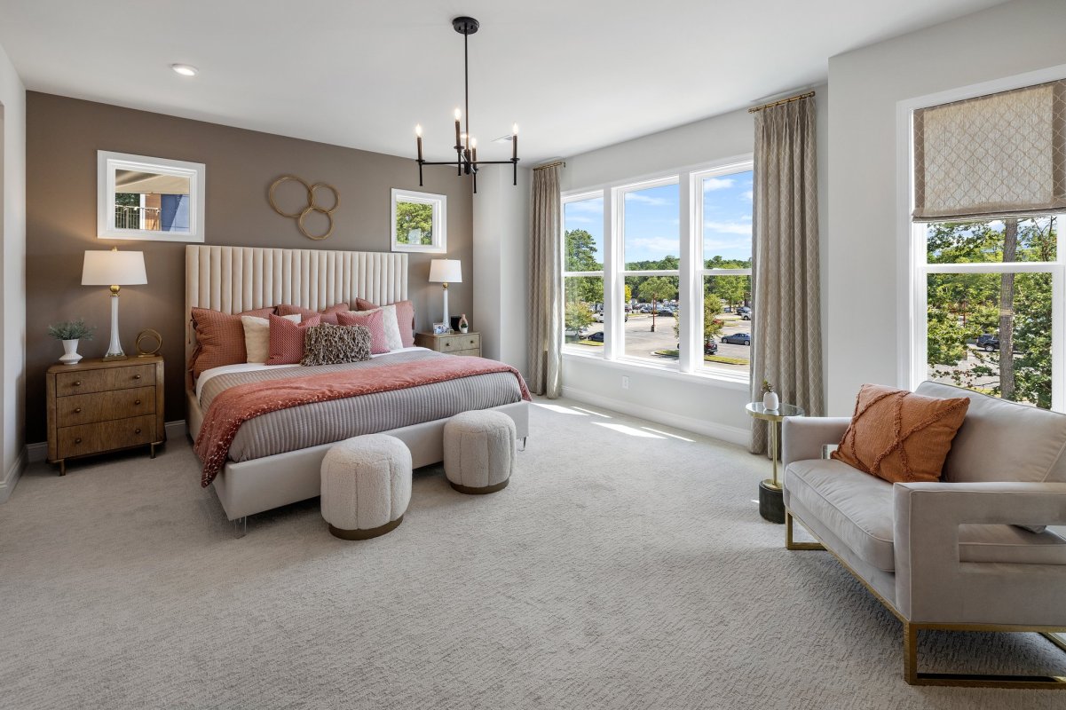 Spacious primary suite in the Drake floor plan at The Aire At Westchester by HHHunt Homes, featuring a plush upholstered bed with accent pillows, two nightstands with lamps, a cozy seating area, large windows with abundant natural light, and neutral-toned