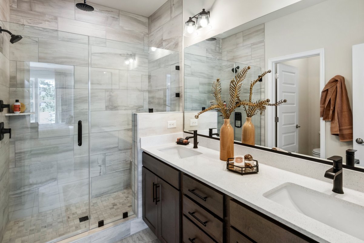 Elegant primary bathroom in the Drake floor plan at The Aire At Westchester by HHHunt Homes, featuring a double vanity with quartz countertops, matte black fixtures, decorative lighting, and a large walk-in shower with glass doors and marble-style tile.