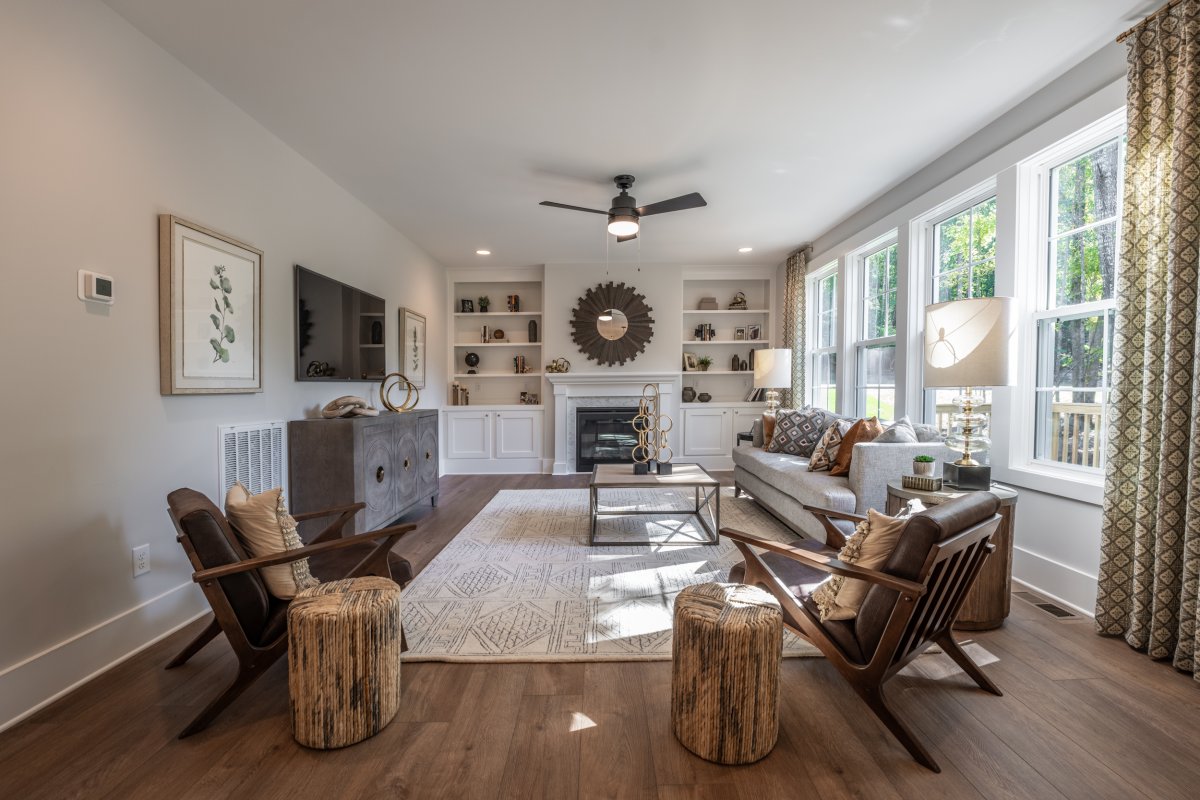 Bright and inviting living room in the Grayson floor plan at Bethesda Forest by HHHunt Homes, featuring a fireplace with built-in shelves, large windows with patterned curtains, modern accent furniture, and wood-style flooring with a neutral area rug.