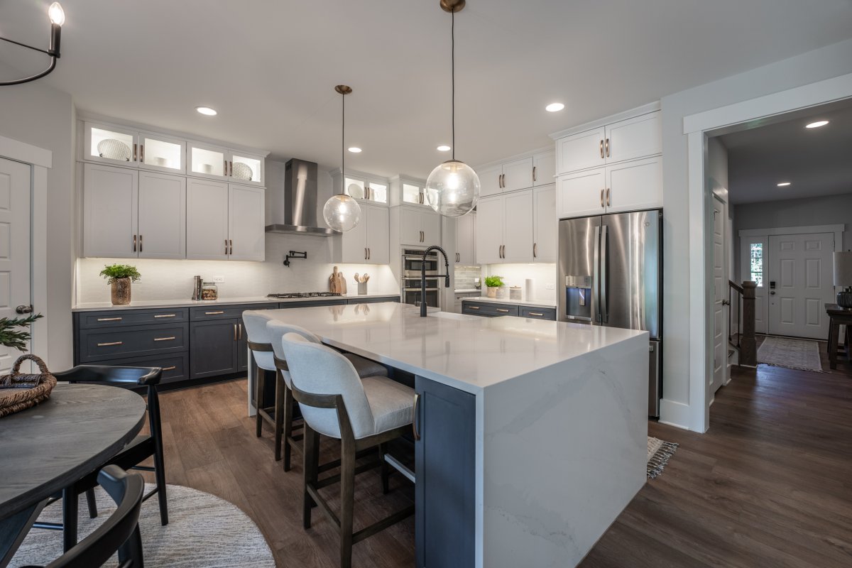 Gourmet kitchen in the Grayson floor plan at Bethesda Forest by HHHunt Homes, featuring a large waterfall-edge island with quartz countertops, two-tone cabinetry in navy and white, stainless steel appliances, globe pendant lights, and wide-plank hardwood 
