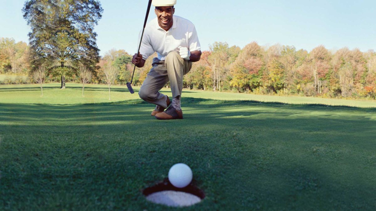 Smiling golfer celebrating a successful putt on a green fairway, with a golf ball near the hole and colorful autumn trees in the background under a clear blue sky.