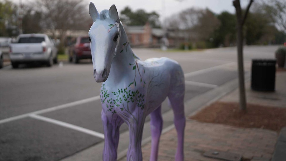 Painted horse statue in downtown Southern Pines, NC, featuring a whimsical tree and leaf design in shades of lavender and green, placed on a brick sidewalk along a quiet street with parked cars in the background.