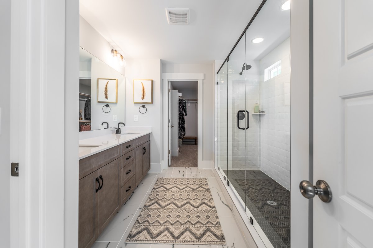 Modern primary bathroom in the Grayson floor plan at Bethesda Forest by HHHunt Homes, featuring a double-sink vanity with quartz countertops, matte black fixtures, a large walk-in shower with frameless glass doors and white tile walls, and access to a wal