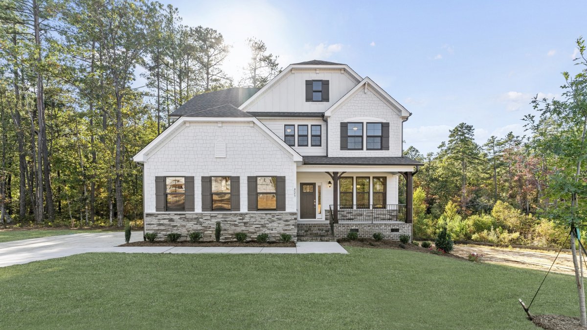 Exterior view of the Grayson European C elevation at Bethesda Forest by HHHunt Homes, featuring a two-story home with white siding, dark shutters, stone accents, a covered front porch with wood columns, and a large landscaped front yard surrounded by tree