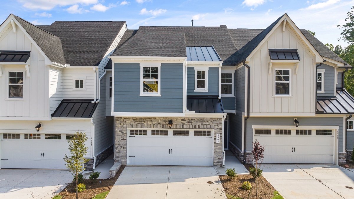 Exterior view of Mount Hermon townhomes by HHHunt Homes featuring a two-story design with blue and beige siding, white trim, black metal roof accents, and two-car garages.