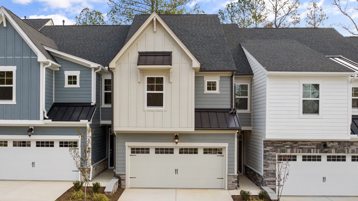 Exterior view of a modern two-story townhome by HHHunt Homes at Mount Hermon, featuring a two-car garage, mixed siding in beige and blue with board-and-batten accents, black metal awnings, and landscaped front entry.-