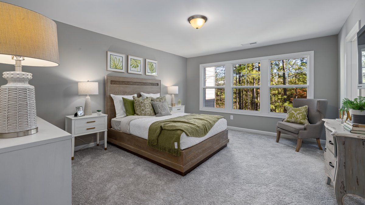 Bedroom in a Mount Hermon townhome by HHHunt Homes featuring a large window wall with wooded views, upholstered bed with green and white bedding, matching nightstands with lamps, and plush carpet flooring.