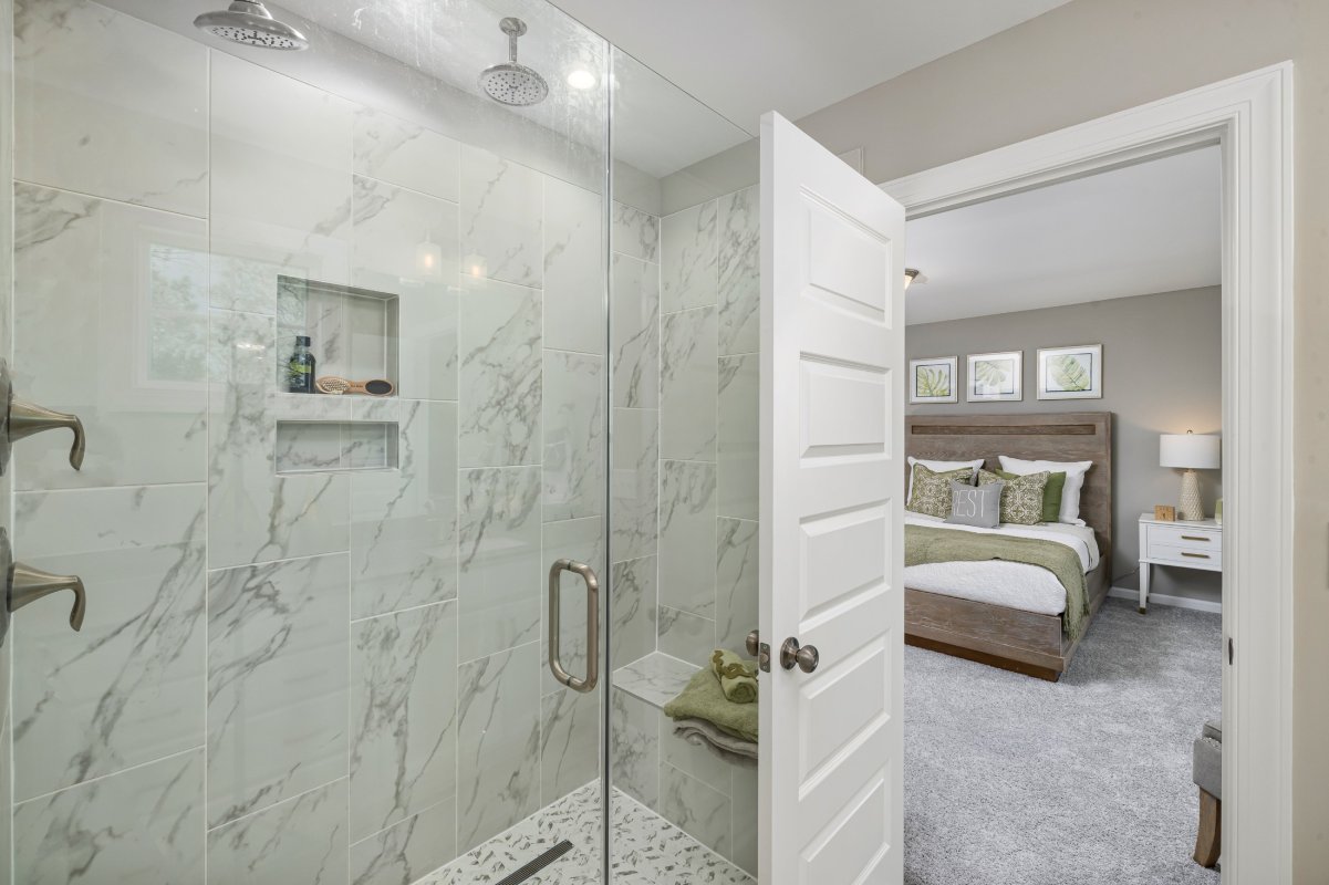 Bathroom in a Mount Hermon townhome by HHHunt Homes featuring a glass-enclosed walk-in shower with marble-look tile, built-in niche, and rainfall showerhead, leading into a carpeted bedroom with modern decor.