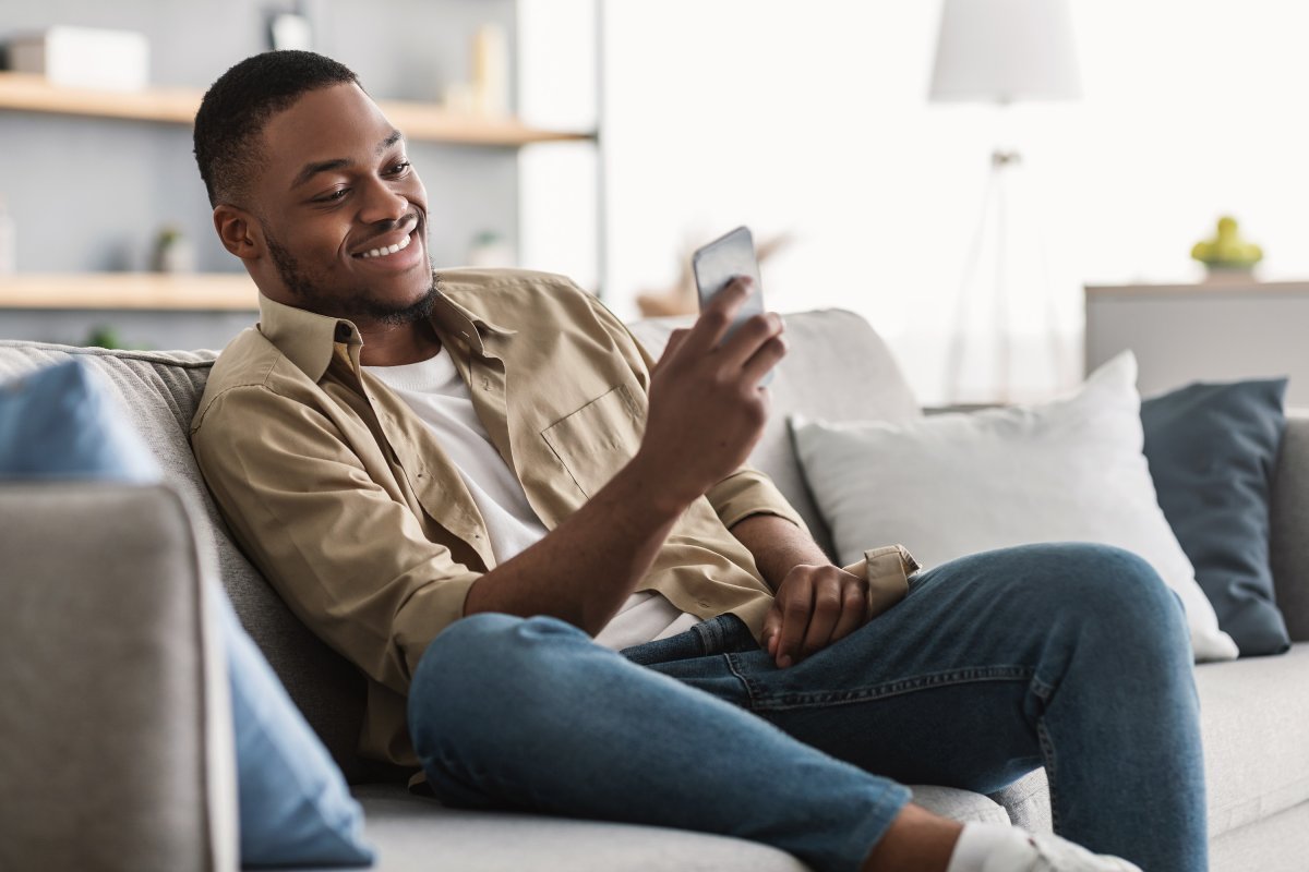 A man sitting on the couch in his new home, smiling and looking at his phone