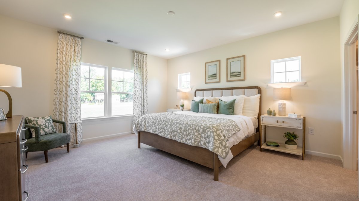 Spacious primary suite in the Maxwell  floor plan by HHHunt Homes at Oak Meadow, featuring a tufted upholstered bed, matching nightstands with lamps, and soft carpeting